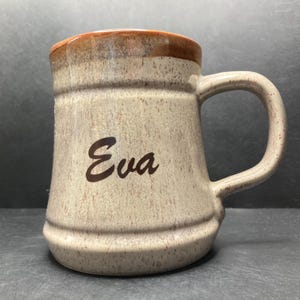 May include: A beige ceramic mug with a brown rim and a brown speckled glaze. The mug has the name "Eva" written on the front in brown lettering.