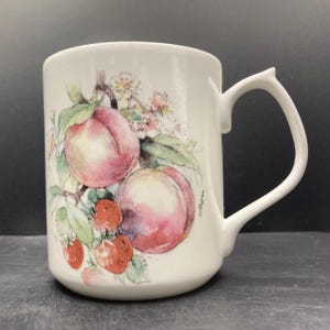 May include: White ceramic mug featuring a watercolor illustration of peaches, strawberries, and blossoms. The handle has a unique, curved design. The mug is a standard size for coffee or tea.