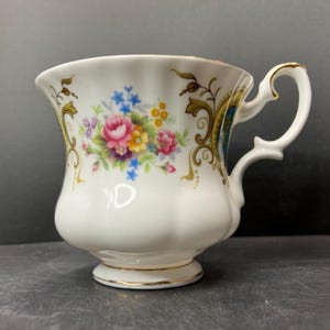 Könnte beinhalten: Weiße Teetasse mit floralem Design mit rosa Rosen, blauen und gelben Blumen und goldenen Akzenten. Der Griff und der Sockel sind ebenfalls mit Gold verziert. Die Tasse hat einen gewellten Rand und einen Sockel.