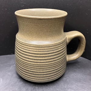 May include: A stoneware mug in a muted olive green hue. The mug features a cylindrical body with horizontal ridged detailing and a curved handle. The top flares slightly outward. The mug is suitable for hot or cold beverages.