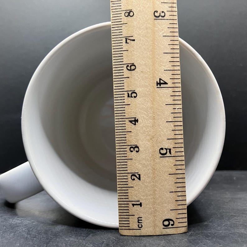 Puede incluir: Una taza de cer&aacute;mica blanca con asa, vista desde arriba. Una regla de madera se coloca dentro de la taza, mostrando una medida de aproximadamente 8 cm. La taza est&aacute; sobre una superficie oscura.
