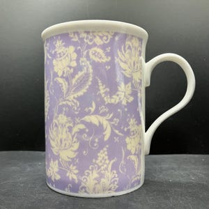 May include: A ceramic mug with a white handle and rim. The mug has a lavender background with a cream floral design. The cylindrical mug is suitable for hot or cold drinks.