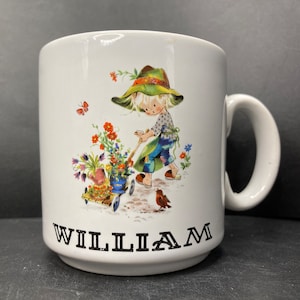 Könnte beinhalten: Weiße Keramiktasse mit einer bunten Illustration eines Mädchens, das eine Schubkarre voller Blumen schiebt. Das Mädchen trägt einen grünen Hut und eine blaue Latzhose. Die Tasse trägt den Namen "WILLIAM" auf dem Boden.