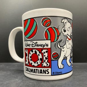 Op de afbeelding: Witte keramische mok met een ontwerp van Disney's 101 Dalmatiërs. De mok heeft een wit handvat en een rondom grafische afbeelding van een Dalmatische hond, rood, turquoise en wit gestreepte ballen en het Disney-logo.