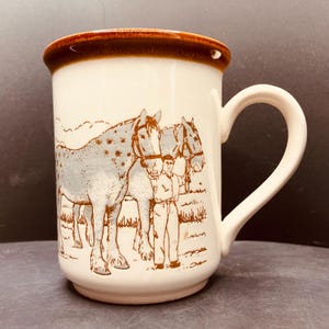 May include: A cream-coloured ceramic mug featuring a brown rim and a brown graphic illustration of a person standing between two draft horses. The horses have spotted coats and are wearing bridles. The mug has a curved handle on the right side.