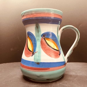Op de afbeelding: Een keramische kan met een witte basis en horizontale banden in turquoise, blauw en roodbruin. Het lichaam is versierd met twee grote, gestileerde oogvormige motieven in blauw, roodbruin en geel. De kan heeft een gebogen handvat aan de rechterkant.