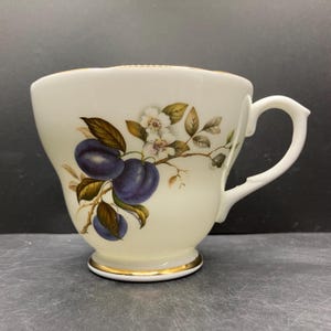 May include: White teacup with a gold rim and handle, featuring a plum and floral design. The teacup has a curved shape and a decorative base. The plums are a deep purple color, and the flowers are white with yellow centers.