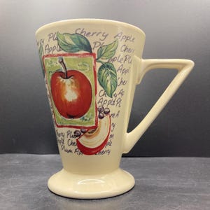 May include: Cream-coloured ceramic mug with a square handle and a pedestal base. The mug features a red apple illustration with green leaves and the words "Cherry Apple Plum" in a handwritten style. The mug is approximately 15 cm tall.
