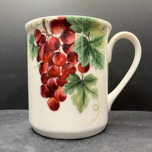 Könnte beinhalten: Weißer Keramikbecher mit einer lebendigen Illustration einer Traube mit roten Trauben und grünen Blättern. Der Becher hat einen gebogenen Griff und einen leicht ausgestellten Rand. Das Design ist auf der Außenseite des Bechers zentriert und bietet eine klassische, fruchtige Ästhetik.