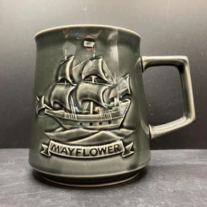 May include: A gray ceramic mug with a square handle. The mug features a raised relief of a sailing ship with the word "MAYFLOWER" on a banner below the ship. The mug has a glossy finish.