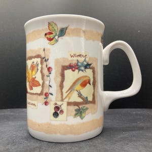 Op de afbeelding: Witte keramische mok met seizoensgebonden illustraties. De mok heeft een oor en is versierd met afbeeldingen van herfstbladeren, bessen en een vogel, met het woord "Winter" zichtbaar. Beige strepen omcirkelen de mok.