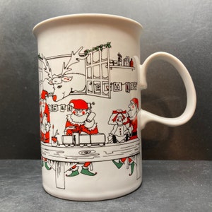 May include: White ceramic mug with a Christmas scene featuring Santa Claus, elves, and a reindeer. The mug has a red handle and a black outline around the design.