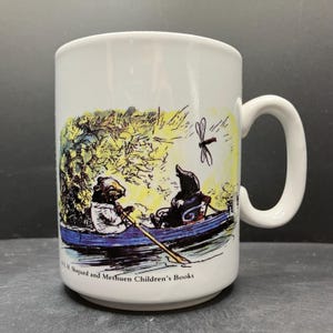 May include: White ceramic mug featuring a whimsical illustration of two animal characters in a blue rowboat, with a dragonfly overhead. The mug has a curved handle and the text "E.H. Shepard and Methuen Children's Books" at the bottom.