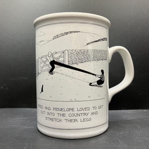 Peut inclure: Mug en céramique blanche avec une illustration en noir et blanc. L'illustration représente deux personnages s'étirant les jambes dans un cadre rural. Le texte en bas dit : "Fred et Pénélope aimaient sortir à la campagne et s'étirer les jambes."