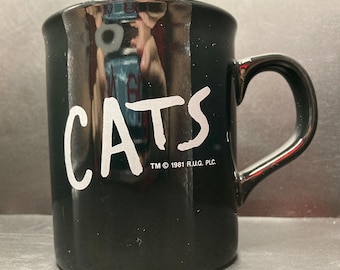 May include: Black ceramic mug with the word "CATS" in white lettering. The mug has a curved handle and the text "TM © 1981 R.U.G. PLC." is printed below the word "CATS".