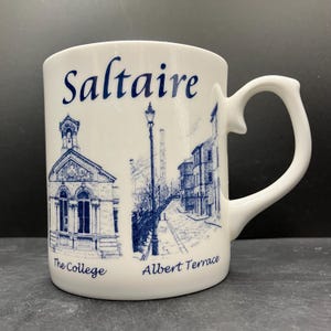 May include: White ceramic mug featuring a blue illustration of Saltaire, with the words "Saltaire", "The College", and "Albert Terrace". The mug has a unique handle design and is suitable for hot or cold beverages.