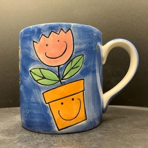 Op de afbeelding: Een keramische mok met een blauwe achtergrond en een grillig bloemmotief. De bloem is roze met een lachend gezicht, groene bladeren en is geplant in een oranje pot met een lachend gezicht. De handgreep is wit.