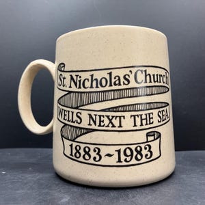 May include: A beige ceramic mug with a speckled finish. The mug features a black banner design with the text "St. Nicholas' Church WELLS NEXT THE SEA 1883~1983". The mug has a curved handle.