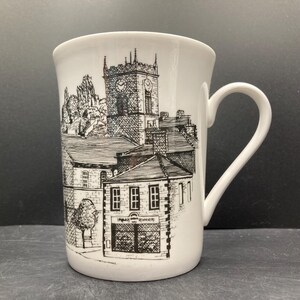 May include: White ceramic mug featuring a black and white illustration of a town scene with a church tower and buildings. The mug has a curved handle and a slightly flared rim. The illustration includes the text "TABAK & RONGERIE".