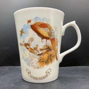 May include: White ceramic mug featuring a detailed illustration of pheasants in flight and perched on a rock. The birds have vibrant orange, brown, and blue plumage. The mug has a curved handle and the word "Sandringham" is printed on the bottom.