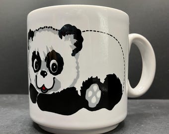 May include: White ceramic mug featuring a cartoon panda bear design. The panda has black and gray accents, with a smiling face and a red mouth. The mug has a curved handle and is suitable for hot or cold beverages.