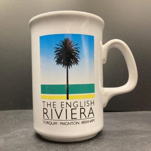May include: White ceramic mug with a handle. The mug features a design of a palm tree under a blue sky, with green and yellow stripes. Text on the mug reads "THE ENGLISH RIVIERA" and lists Torquay, Paignton, and Brixham.