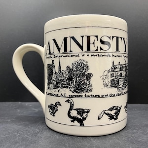May include: Cream-colored ceramic mug with a black graphic design. The design includes the word "AMNESTY" and the text "Amnesty International is a worldwide human rights". The mug also features illustrations of buildings and geese.