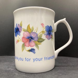 May include: White ceramic mug with a floral design featuring blue, pink, and purple pansies and green leaves. The mug has a curved handle and the words "thank you for your friendship" in blue.