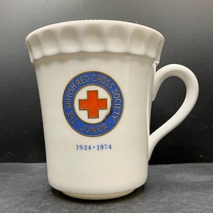Op de afbeelding: Witte keramische mok met een geschulpte rand en een gebogen oor. De mok heeft een cirkelvormig embleem met de tekst "THE BRITISH RED CROSS SOCIETY JUNIOR" rond een rood kruis. De jaartallen "1924 1974" staan eronder.