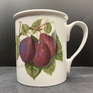 May include: A white ceramic mug featuring a botanical illustration of three dark purple plums on a branch with green leaves. The text "Reine Claude Plum" is printed in small black script near the base of the fruit illustration on the mug.