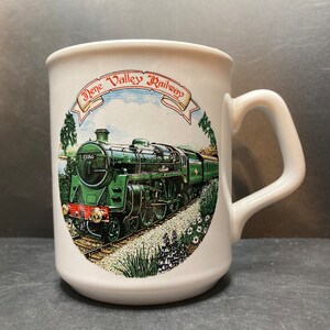 May include: White ceramic mug with a green, black, and red steam train on it. The train is surrounded by green foliage and flowers. The text "Dene Valley Railway" is printed in red on the mug.