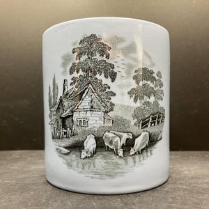 May include: A white ceramic mug with a black and white pastoral scene. The design features a cottage, trees, and cows near a body of water. The mug has a cylindrical shape and a smooth surface.