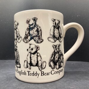 Peut inclure: Mug en céramique blanche orné d'illustrations de nounours noirs et blancs. La tasse a une anse incurvée et le texte "The English Teddy Bear Company" est imprimé sur le côté.