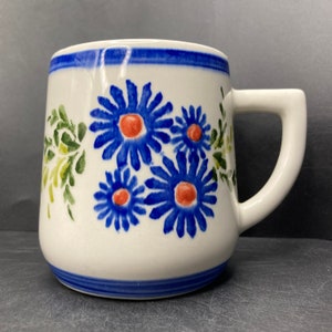 May include: White ceramic mug featuring a blue rim and handle, decorated with blue daisy-like flowers with orange centres. Green leafy accents enhance the floral design. The mug has a sturdy, square handle.