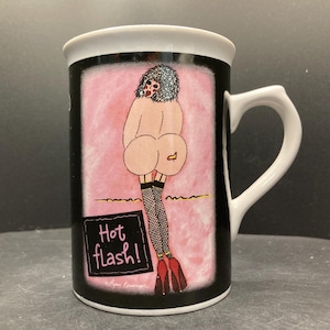Op de afbeelding: Witte keramische mok met een zwarte achtergrond en een roze midden. De mok heeft een cartoonillustratie van een persoon van achteren, met visnetkousen en rode hakken. De tekst "Hot flash!" wordt op de mok weergegeven.
