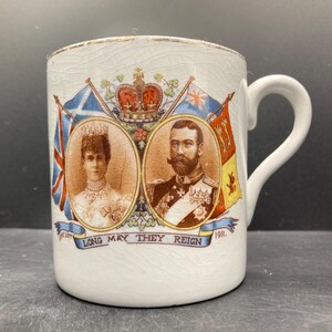May include: White ceramic mug featuring portraits of a royal couple, flags, and a crown. The mug has the text "LONG MAY THEY REIGN 1911." The mug has a white handle and a gold rim. The mug is a commemorative item.
