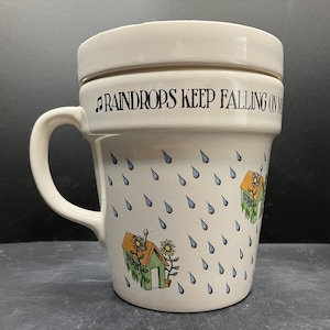 May include: Cream-colored ceramic mug with a handle and a lid. The mug is decorated with blue raindrops and small green houses with orange roofs and yellow flowers. The text "Raindrops Keep Falling On" is printed on the mug.
