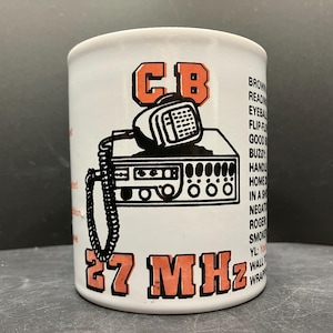 May include: White ceramic mug featuring a black and orange graphic of a CB radio with a microphone and the text "CB" and "27 MHz". The mug also has a list of CB radio terms printed on the side.