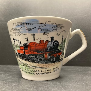 May include: A white ceramic teacup with a red, black, and gray steam engine train illustration. The text on the cup reads "Ex. B.R. Class 2. 2.6.0. 6441 At Steam Town, Carnforth, Lancs."