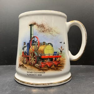 May include: White ceramic mug with a gold rim and handle. The mug features a colorful illustration of a vintage steam-powered rotary cultivator. The text "ROTARY CULTIVATOR by RICKETT 1858" is printed on the mug.