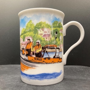 May include: White ceramic mug with a colourful illustration of an orange lifeboat with two figures in yellow jackets, the text "R.N.L.I.", and a coastal town scene. The mug has a curved handle and a blue and white wave design at the bottom.