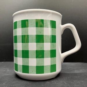 May include: White ceramic mug with a green and white checkered pattern. The mug has a curved handle and a cylindrical shape. The checkered design covers the entire body of the mug, creating a classic, retro aesthetic.