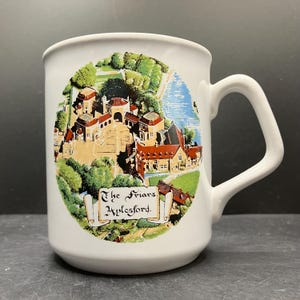 May include: White ceramic mug featuring a colorful illustration of a village scene with red-roofed buildings, trees, and a river. The text "The Friars, Aylesford" is displayed on a scroll. The mug has a large handle.