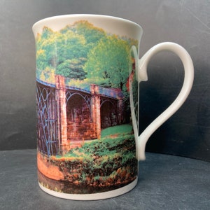 May include: White ceramic mug with a colorful image of a stone bridge with blue metal supports over a canal. The image is surrounded by green trees and foliage.
