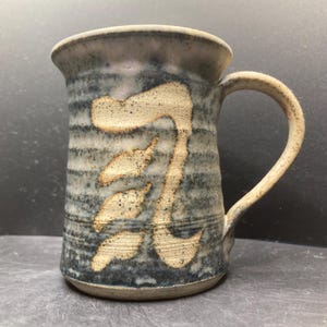 Op de afbeelding: Een handgemaakte keramische mok met een beige handvat en een cilindrische body. De mok heeft een getextureerde, blauwe en grijze buitenkant met een beige, gestileerd karakterontwerp. De mok is geschikt voor warme of koude dranken.
