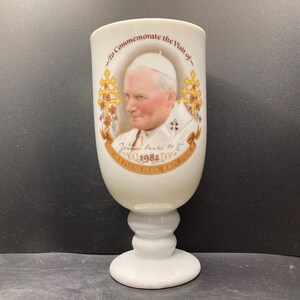 May include: White commemorative goblet featuring a portrait of Pope John Paul II, with text that reads "To Commemorate the Visit of - His Holiness Pope John Paul II 1982". The goblet has a bulbous body and a pedestal base.