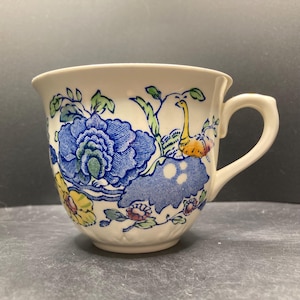 Op de afbeelding: Crèmekleurige theekop met een bloemmotief. De kop heeft een grote blauwe bloem, gele en roze accenten en groene bladeren. Het handvat is gebogen en de kop heeft een licht uitlopende rand. Het ontwerp doet denken aan vintage servies.