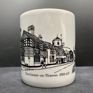 May include: White ceramic mug featuring a black and white illustration of a street scene with buildings and the text "Dorchester-on-Thames 2000 A.D." The mug has a cylindrical shape.