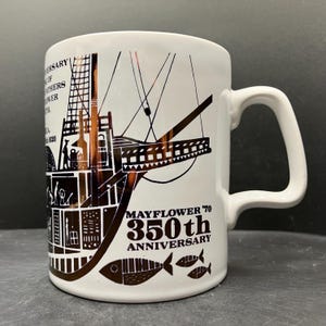 May include: White ceramic mug featuring a brown and black illustration of the Mayflower ship. The mug commemorates the 350th anniversary of the Mayflower voyage, with text and fish illustrations. The mug has a white handle.
