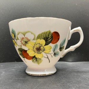 Könnte beinhalten: Weiße Teetasse mit floralem Design mit gelben und weißen Blumen, grünen Blättern und roten Beeren. Die Tasse hat einen goldfarbenen Griff und Sockel, und das Blumenmuster befindet sich außen.